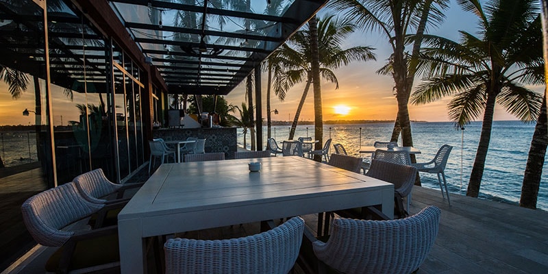 An outdoor dining area is set against the backdrop of a sunset over the water. The space features a large white table surrounded by several wicker chairs. Palm trees frame the scene, while a clear sky gradually transitions to twilight.