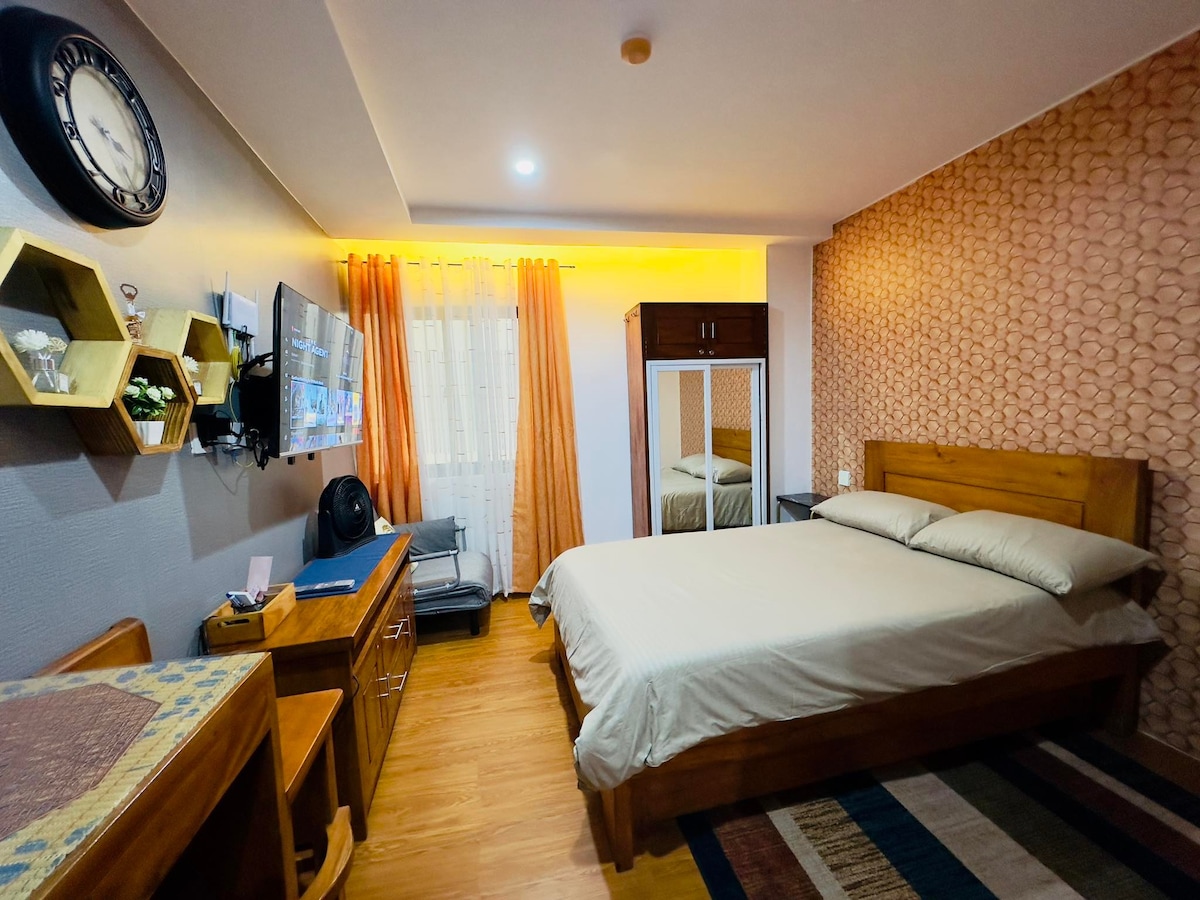 The studio unit features a spacious bed with neutral bedding against a textured accent wall. A television is mounted on the wall opposite, with a wooden desk and shelves nearby. Natural light filters through sheer curtains, complementing the warm tones of the room.