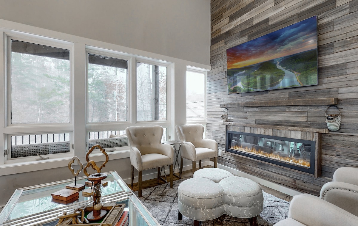 The living area features a modern design with a stone accent wall and a 65-inch television mounted above a sleek electric fireplace. Two plush armchairs provide seating next to a stylish coffee table, while large windows allow natural light to fill the space.