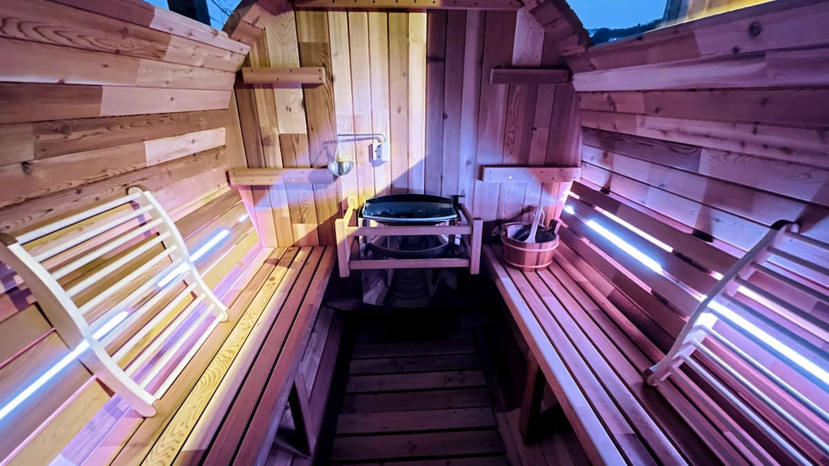 The interior of a Finnish barrel sauna is showcased, featuring warm wooden paneling and benches for seating. Ambient lighting illuminates the space, enhancing the wooden textures. A heater is positioned centrally, complementing the serene atmosphere suitable for private relaxation.