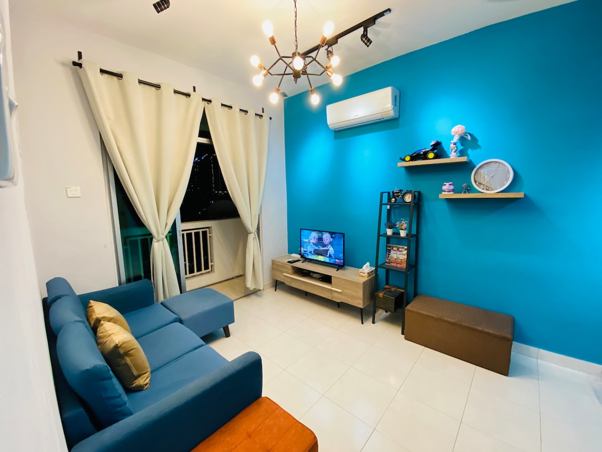 A living area is presented with a blue accent wall and modern light fixtures. A comfortable blue sofa and an orange ottoman are positioned near a media console, which holds a television. Natural light filters in through white curtains covering a balcony door.