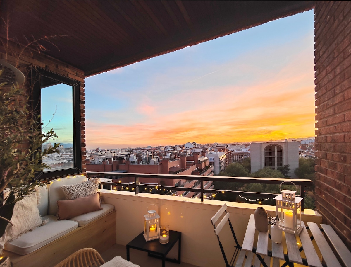 A spacious terrace showcases stunning skyline views during sunset, framed by warm orange and pink hues. Comfortable seating is arranged alongside a small table, creating an inviting space for relaxation. Soft lighting from lanterns adds a cozy ambiance to the open-air setting.