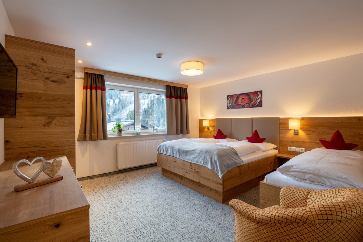 A spacious sleeping area features two wooden beds, each adorned with red decorative pillows. A TV mounted on the wall is visible across from the beds. Natural light brightens the room through a large window, with soft curtains framing the picturesque mountain view.