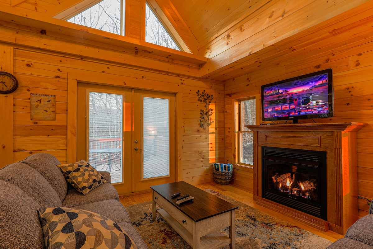 The great room features a gas log fireplace beneath a flat-screen television. Natural light enters through large windows and French doors leading to an outdoor deck. The space includes a comfortable sofa adorned with accent pillows and a wooden coffee table positioned in front.