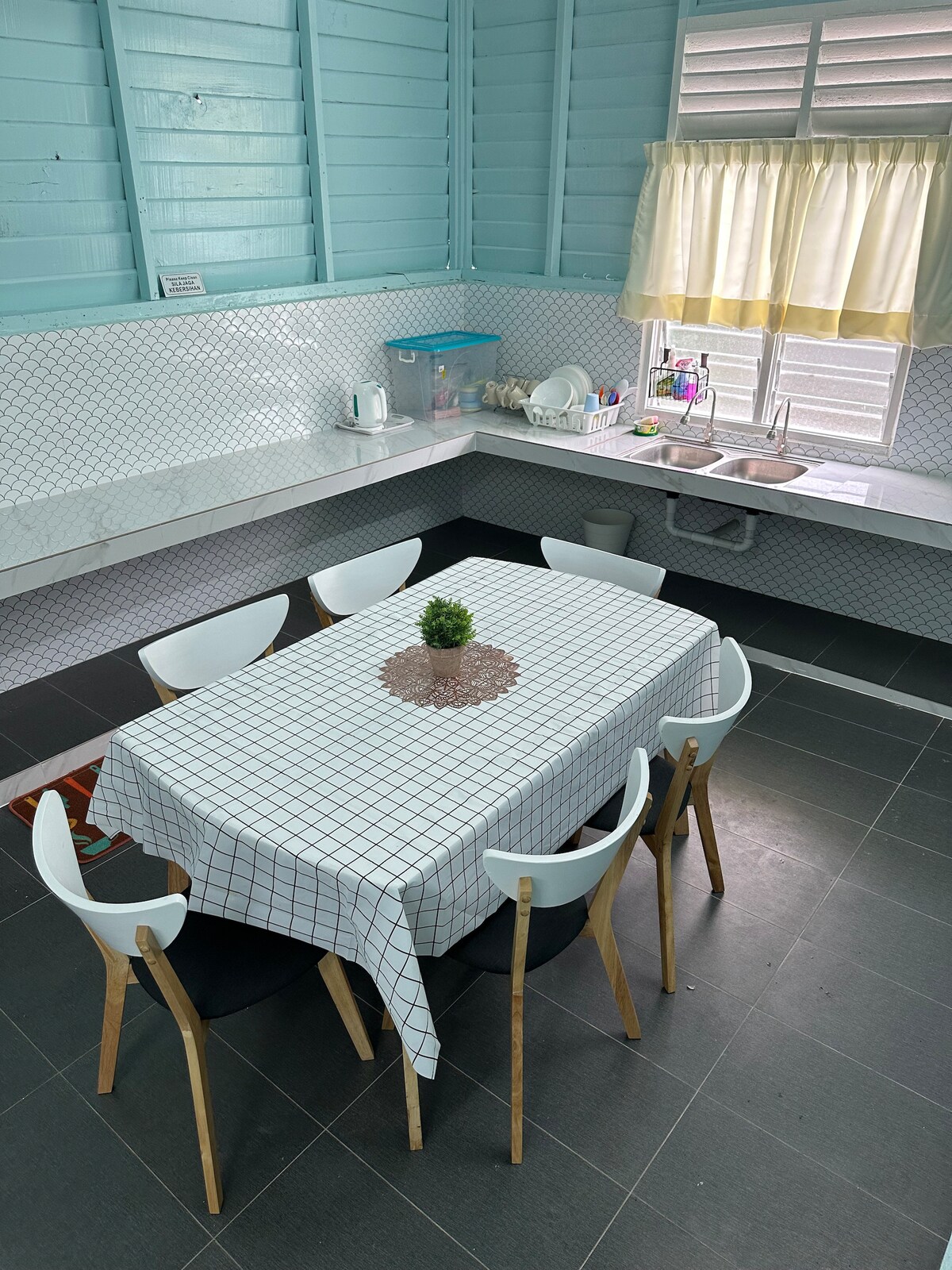 A bright kitchen space features a rectangular dining table surrounded by six white chairs. A simple decorative centerpiece is placed on the table. Natural light enters through a window with sheer curtains, illuminating the countertops and kitchenware arranged nearby.