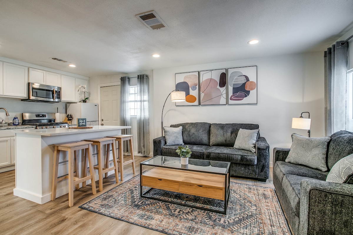 An open living area is shown, featuring a modern kitchen with stainless steel appliances and a spacious breakfast bar with three wooden stools. A comfortable seating arrangement includes a dark gray sofa and a matching loveseat, positioned around a glass coffee table with a wooden base. Decorative artwork adorns the wall.