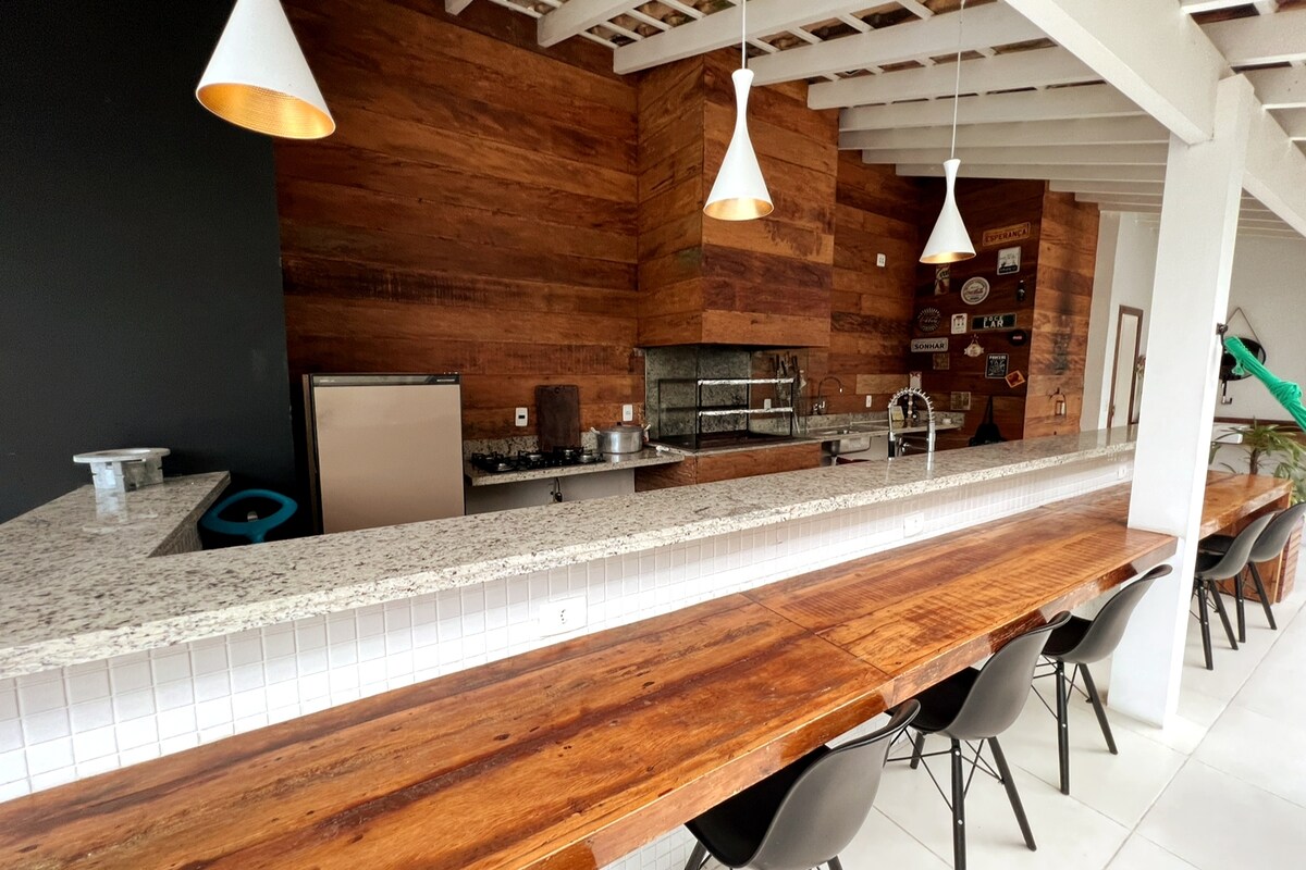 A spacious kitchen area features warm wooden accents on the walls and a large granite countertop. Several pendant lights hang above a long wooden dining table surrounded by a mix of chairs. Stainless steel appliances are positioned for functionality.