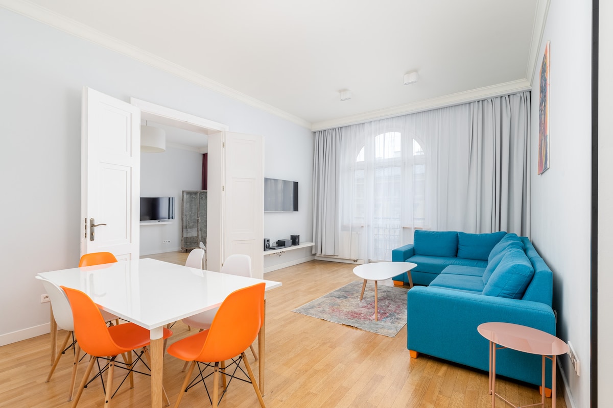 A spacious living area is depicted, featuring a bright blue sofa and a modern dining table surrounded by colorful chairs. Floor-to-ceiling curtains allow natural light to fill the room, highlighting the wooden floor and minimalistic decor. Two televisions are mounted on the wall.