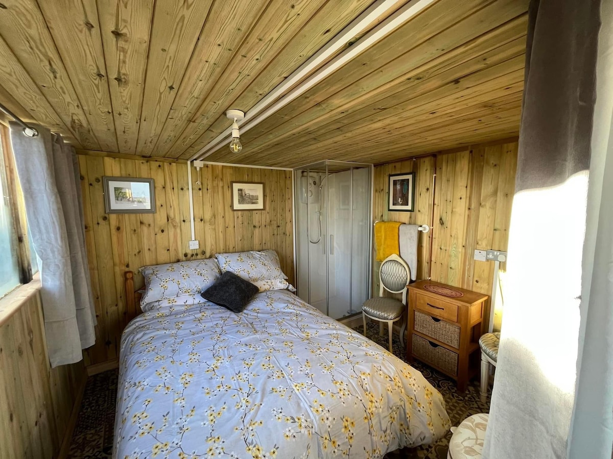 A cozy bedroom with wooden walls and ceiling showcases a spacious bed adorned with a floral comforter. A bedside table and a decorative chair are positioned beside the bed. Light filters through a window, enhancing the inviting ambiance.