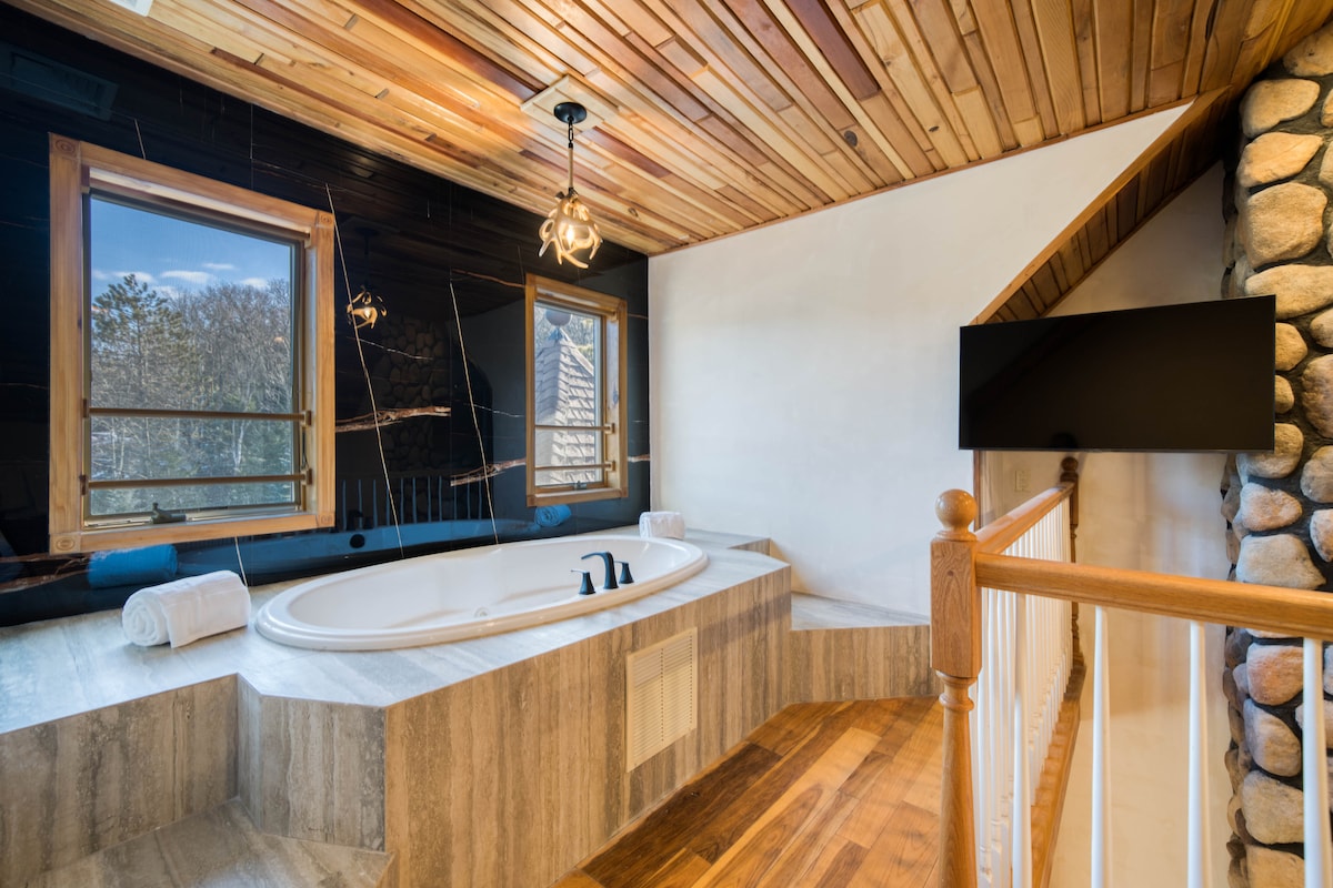 A spacious en-suite bathroom features a large marble Jacuzzi tub set against a backdrop of wooden ceiling beams. Natural light floods the area through two windows, while a sleek television is mounted on the wall. Soft towels are neatly arranged on the ledge.