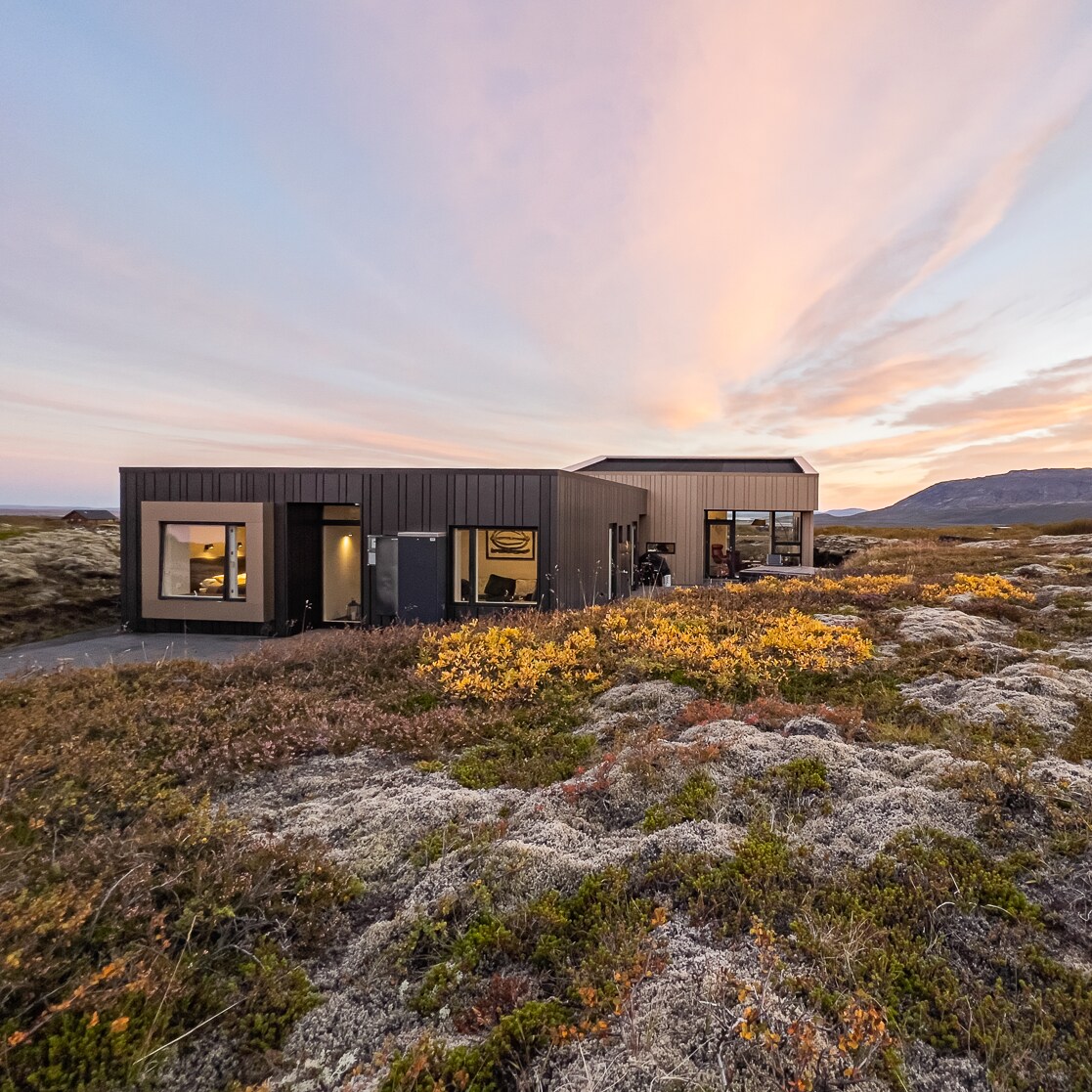 A modern cottage is nestled in a scenic landscape, surrounded by soft moss and low vegetation. Large windows are visible, reflecting the colors of the twilight sky, which transitions from warm pink to cool blue. The structure merges contemporary design with natural beauty.