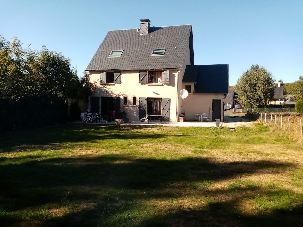 A spacious duplex is depicted, featuring a light-colored exterior with multiple windows. The building is surrounded by a grassy area and trees, creating a natural setting. Outdoor seating is visible, enhancing the space for relaxation or social gatherings.