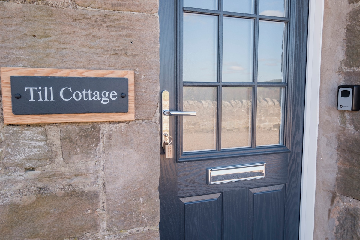 The entrance to Till Cottage features a dark blue door with a modern handle, adorned with a wooden sign displaying 'Till Cottage.' Natural light illuminates the area, enhancing the rustic stone wall providing a welcoming first impression.