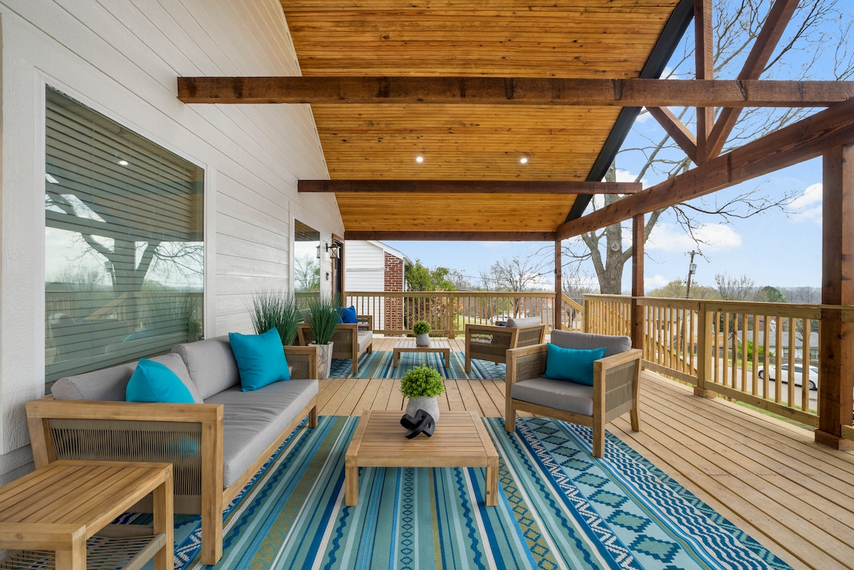 An inviting outdoor seating area features wooden furniture, including a couch and chairs adorned with bright blue cushions. A coffee table sits centrally, surrounded by a patterned area rug. The space is sheltered by a wooden canopy, with a view of greenery in the background.