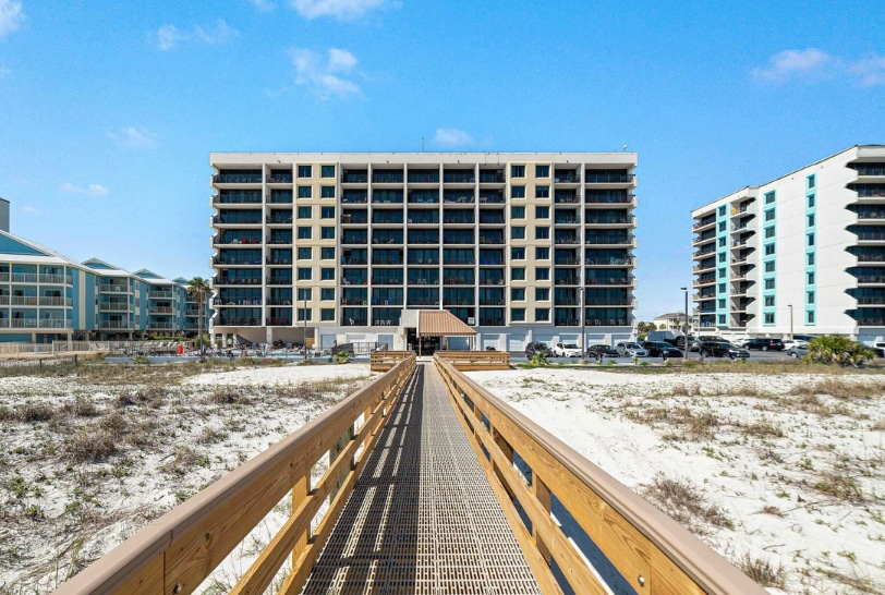 A wooden boardwalk leads to the low-rise condo building nestled along the beach. The structure features multiple floors with several balconies. Soft white sand surrounds the area, providing direct access to the beach, framed by clear blue skies.