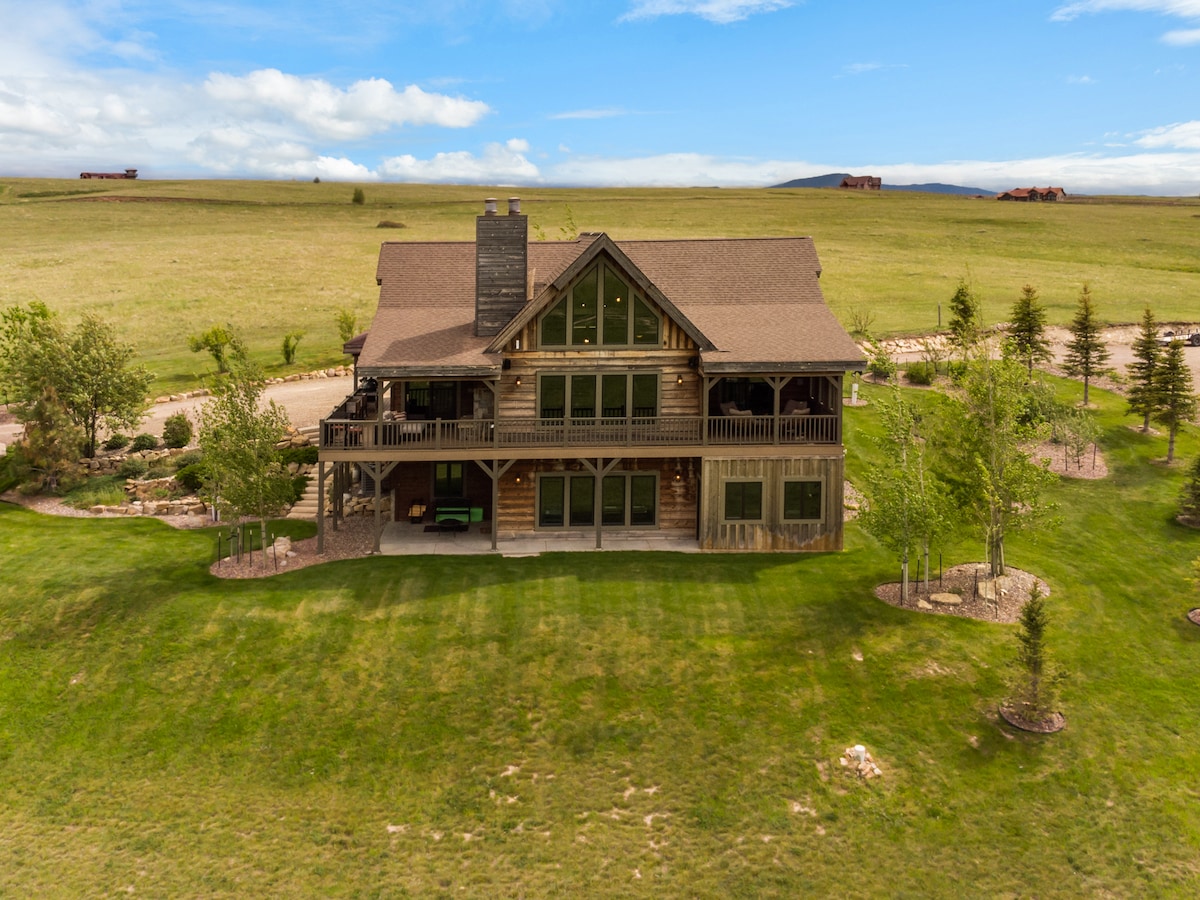 The exterior of the lodge-style home showcases a spacious layout with multiple balconies and large windows. Surrounded by lush green lawns and trees, the structure is situated against a backdrop of rolling hills and a clear blue sky.