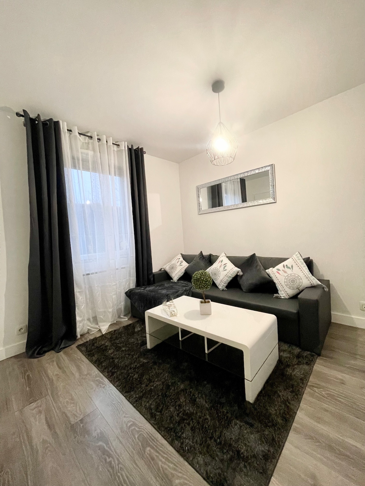 A cozy living area features a dark sofa adorned with decorative pillows. A light-colored coffee table is centered on a plush dark rug, while a mirror reflects natural light from the window draped with sheer and dark curtains.