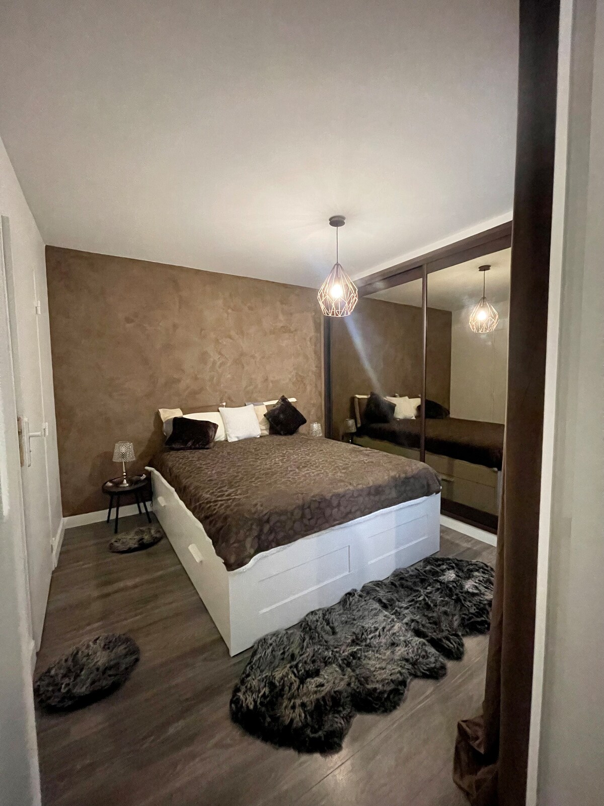 A comfortable bedroom is presented with a spacious bed covered in a dark brown blanket. Soft rugs are placed around the bed, and a closet with mirrored doors creates an illusion of space. A pendant light hangs above, enhancing the warm color of the walls.