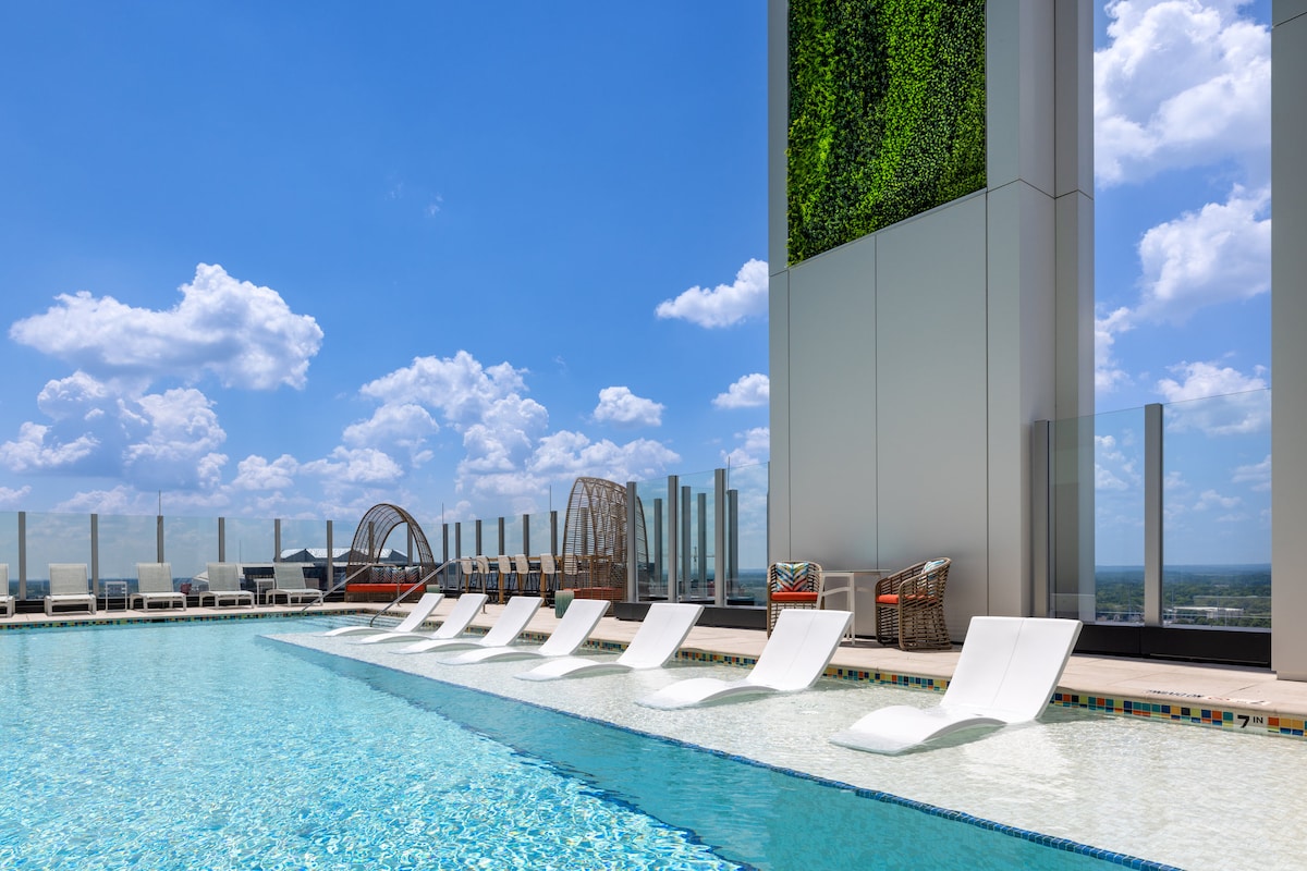 A rooftop pool is visible, featuring a tiled border and several white lounge chairs positioned along the water's edge. A green vertical garden adds a natural element to the modern setting, while expansive city views can be seen in the background under a blue sky with fluffy clouds.