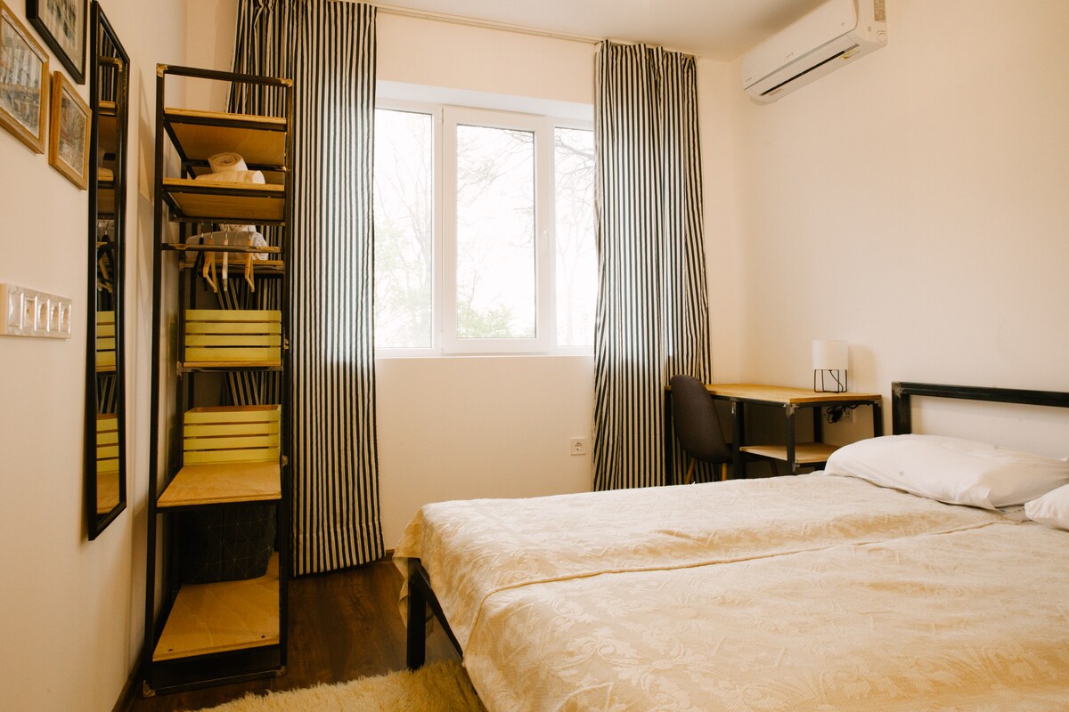 A bright bedroom features a spacious bed with a light-colored duvet. A desk with a chair is set against a wall, alongside a tall shelving unit displaying neatly arranged items. Floor-to-ceiling windows covered by striped curtains allow natural light to fill the room.