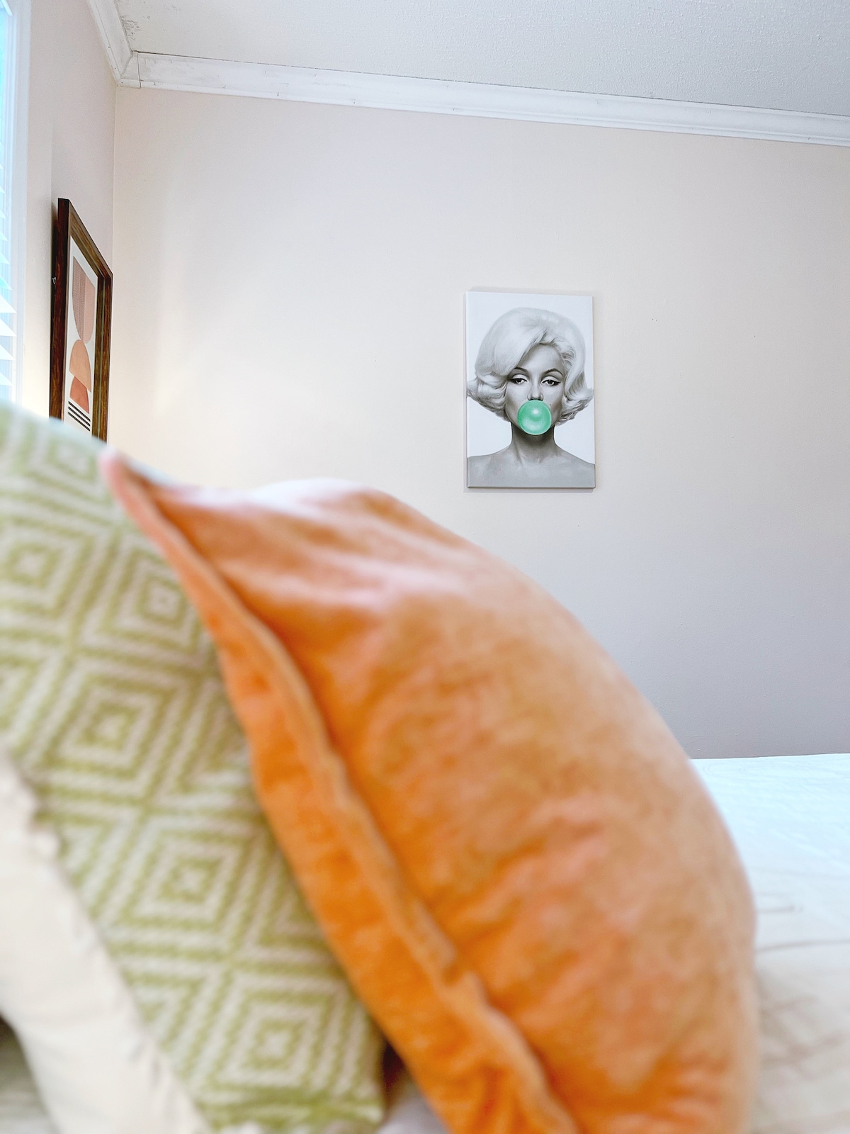 A queen bed is adorned with decorative pillows in shades of orange and green. A framed artwork is displayed on the wall, featuring a unique design. Soft natural light enters through a nearby window, illuminating the calming pastel wall color.