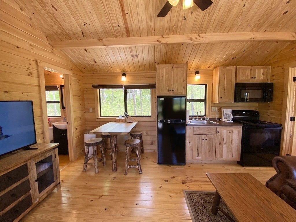 The cabin's interior showcases a warm wooden design with a combined kitchen and living area. A cozy loveseat faces a TV, while a dining table with two stools is positioned near a window. Full-size appliances in the kitchen are visible, creating a functional space.