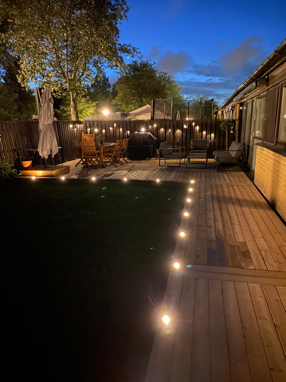 A well-lit outdoor area showcases a wooden deck surrounded by lush greenery. Comfortable seating arrangements are present, along with a dining set. Path lights illuminate the space, creating a welcoming atmosphere for evening gatherings.