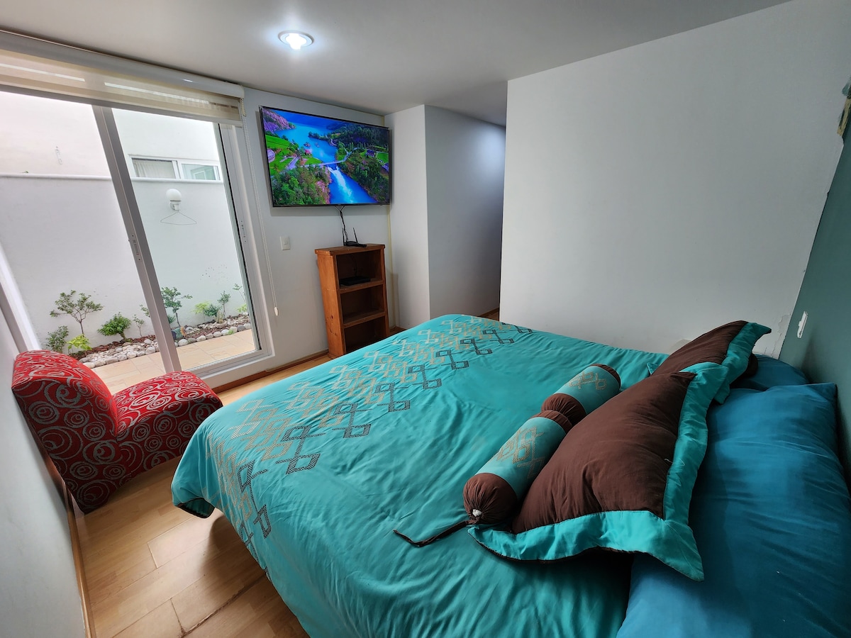 A spacious bedroom is furnished with a large turquoise bedspread, complemented by decorative pillows. A flat-screen TV is mounted on the wall, and a red patterned armchair sits in the corner. Natural light fills the room from the sliding glass doors leading to a private terrace.
