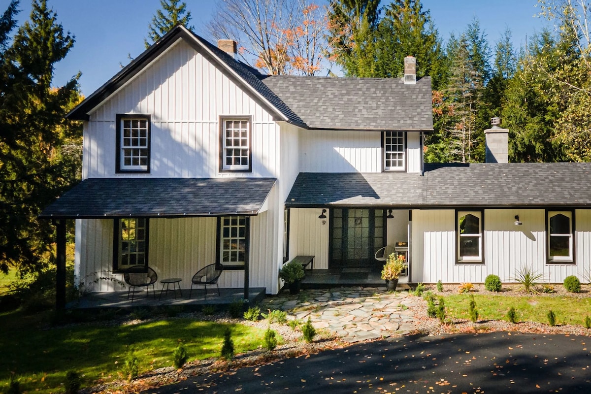 An updated farmhouse exterior features a white façade with black trim and a sloped roof. A covered porch showcases two chairs, while a stone pathway leads to the entrance. Surrounding greenery and trees frame the property, providing a secluded atmosphere.