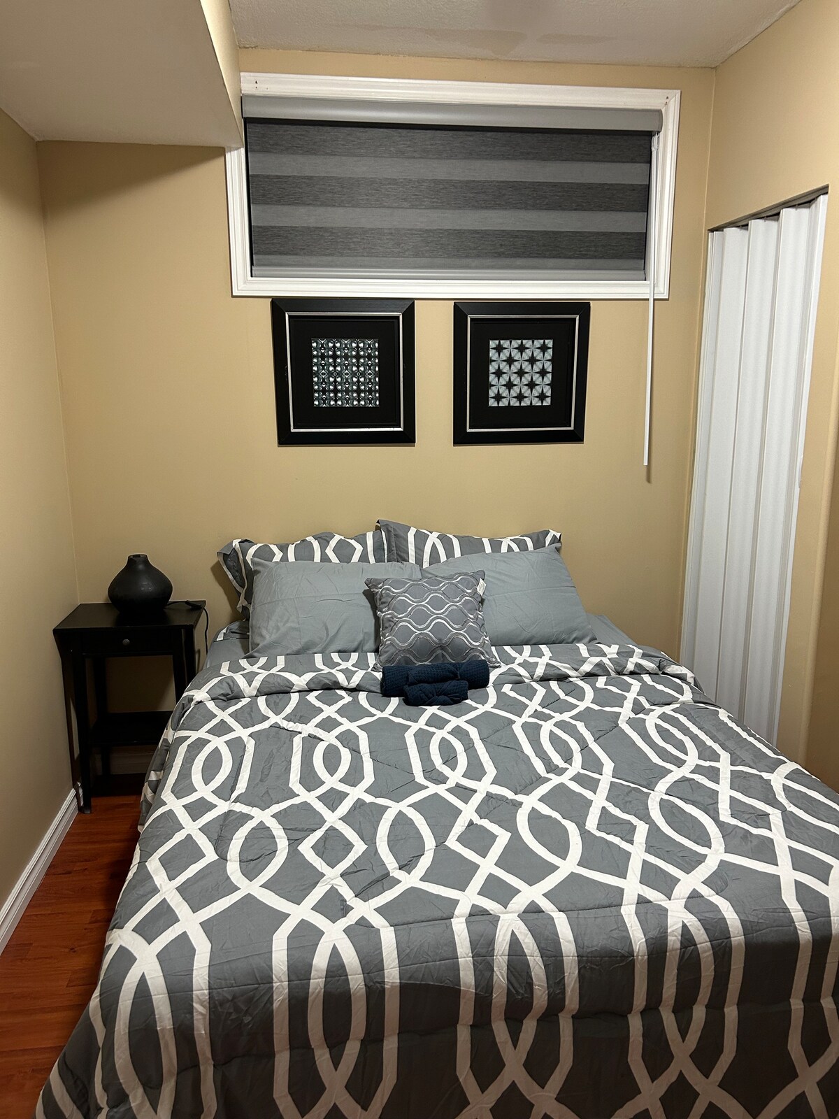 A cozy sleeping area features a neatly made bed with a patterned gray comforter and decorative pillows. Two framed art pieces adorn the wall above the bed. A small nightstand holds a decorative vase, while a window with shades allows soft light to enter the room.