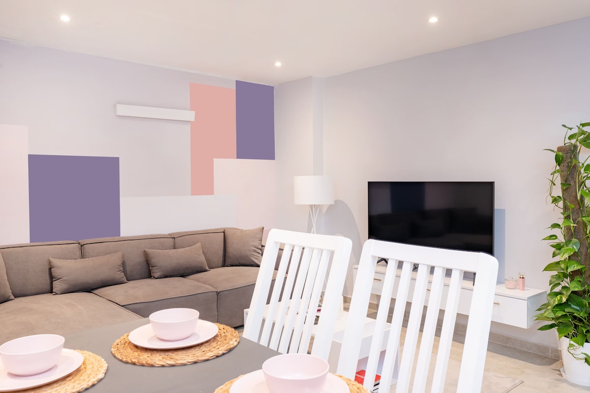 A modern living area is displayed, featuring a spacious gray sectional sofa arranged around a light wood dining table. Overhead lighting creates a bright atmosphere. A large television is mounted on the wall opposite the seating, and a small indoor plant adds a touch of greenery.