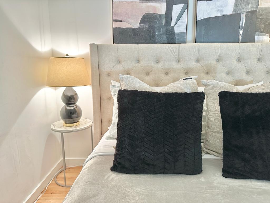 A cozy sleeping area features a tufted headboard alongside decorative pillows in varied textures. A bedside table holds a lamp, providing soft light. Abstract artwork can be seen on the wall, enhancing the room's inviting atmosphere.