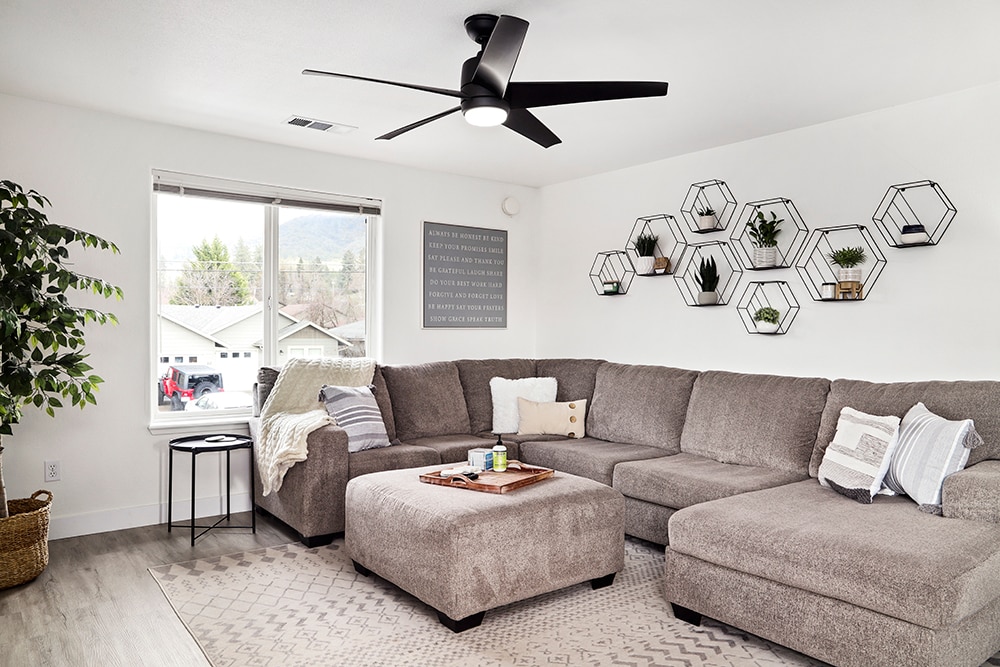 A spacious living area features a large sectional couch arranged for relaxation. A ceiling fan provides airflow, while hexagonal wall shelves display various plants. Natural light enters through a window, complemented by soft decor accents and a neutral color palette.