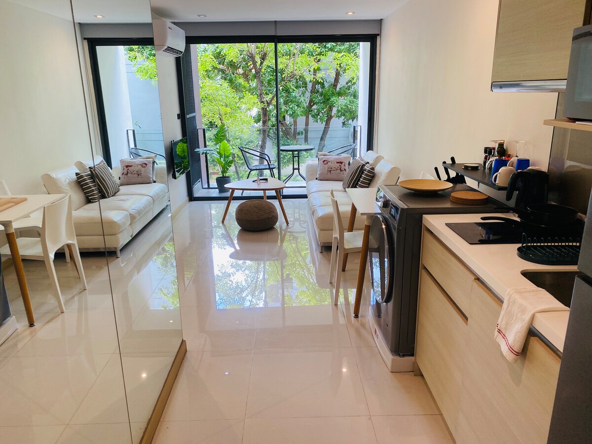 A bright living area features sleek white furniture and a dining table, complementing the open kitchen with modern appliances. A washer is positioned alongside the kitchen, while large glass doors provide access to a balcony overlooking lush greenery.
