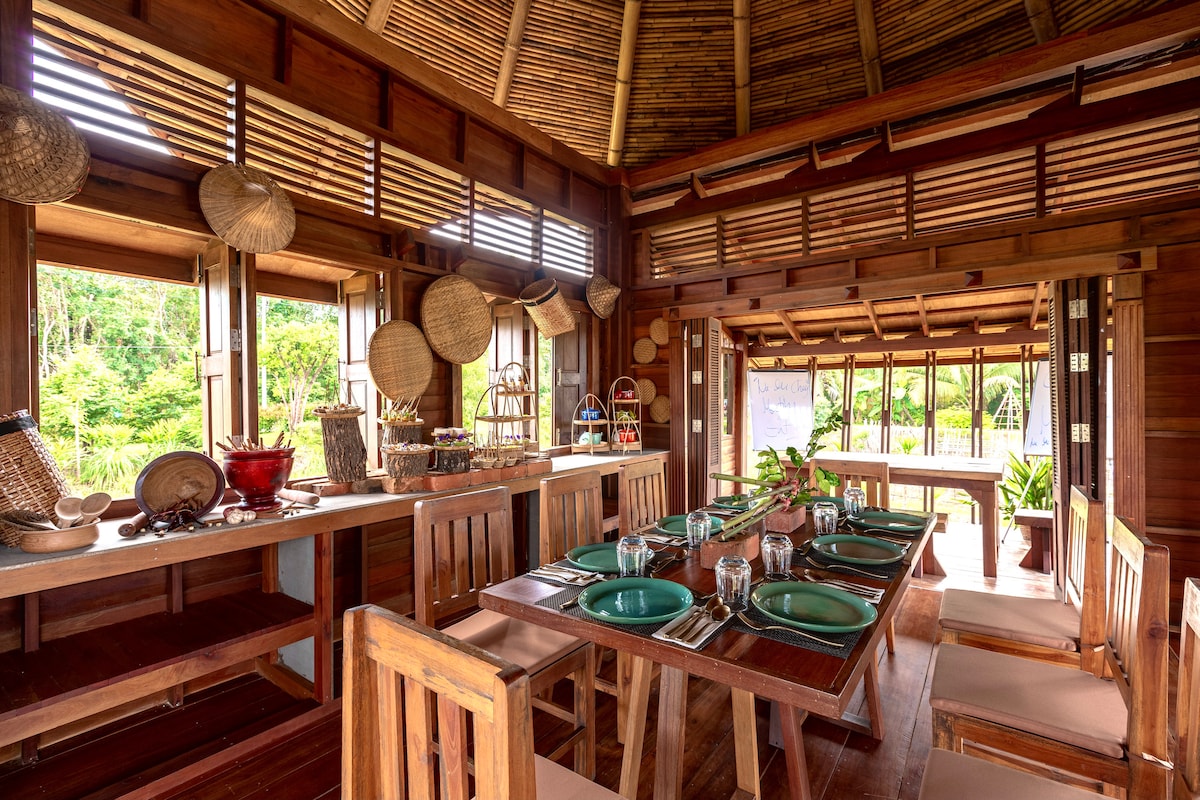 The dining area features wooden tables and chairs arranged around a spacious open layout. Natural light fills the space through large windows, showcasing decorative woven baskets on the walls. A counter displays a selection of items, enhancing the inviting atmosphere of the room.