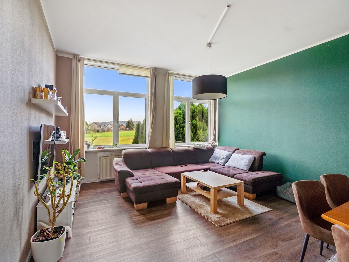 A modern living area features a large sectional sofa with soft cushions, arranged around a wooden coffee table. Natural light fills the space through expansive windows, offering views of greenery outside. A flat-screen TV is mounted on the wall above a minimalist shelf, with tasteful decor nearby.