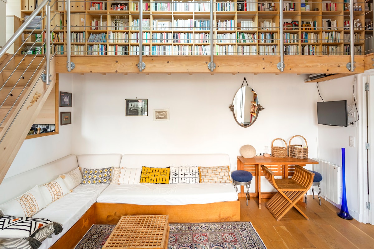 A living space is presented with a light wood floor and a cozy sofa surrounded by patterned pillows. A low coffee table sits atop a decorative rug. A wooden bookshelf filled with books dominates the wall overhead, and a round mirror reflects the room's warmth.