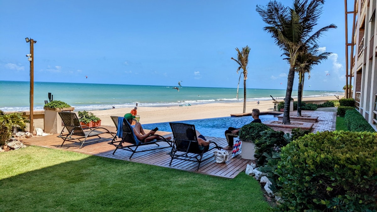 A view of the beachfront shows a tranquil swimming pool adjacent to a wooden deck. Several sun loungers are positioned nearby, facing towards the ocean. Lush greenery surrounds the area, with palm trees swaying gently in the breeze and a clear sky above.
