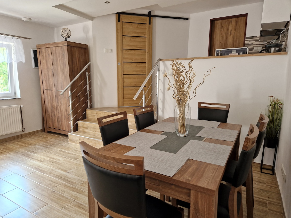 A dining area is presented with a wooden table surrounded by six black chairs. A decorative vase with dried plants is centered on the table. A staircase leads to a higher level, with a wooden door visible at the top. Natural light streams in through the nearby window.