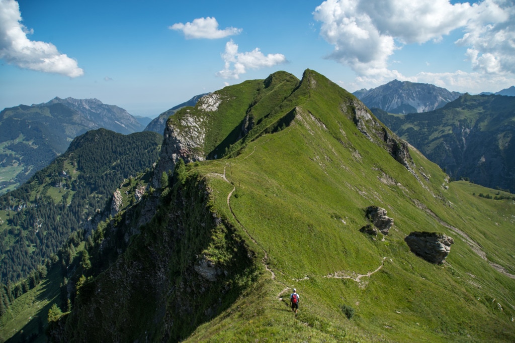 A scenic mountain ridge is displayed, showcasing lush green hills and rocky outcrops under a clear blue sky. A winding path leads along the ridge, with a lone hiker visible in the distance, illustrating the serene beauty of the alpine landscape.