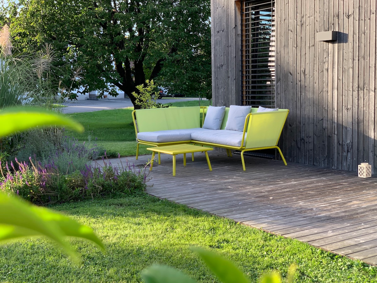 A seating area is presented on a wooden terrace, featuring a modern yellow outdoor sofa with light gray cushions. A small matching table sits in front of the sofa. Lush green grass surrounds the area, complemented by flowering plants in the foreground.