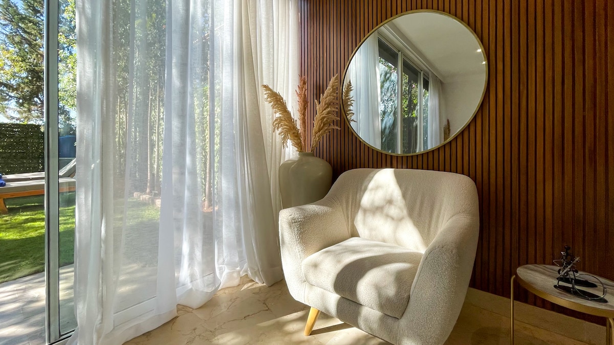 A cozy seating area is highlighted by a plush armchair adorned with light-colored fabric. Natural light floods the space through sheer curtains, casting soft shadows. A round mirror reflects the serene outdoor setting, while a decorative vase with pampas grass adds a touch of elegance.