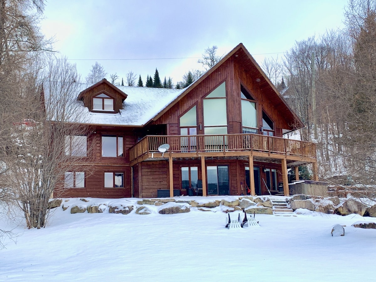 The chalet's exterior is showcased with a wooden facade and a steeply pitched roof. A spacious deck extends from the upper level, offering ample outdoor seating. Snow blankets the ground, and trees are visible in the background, enhancing the serene natural setting.