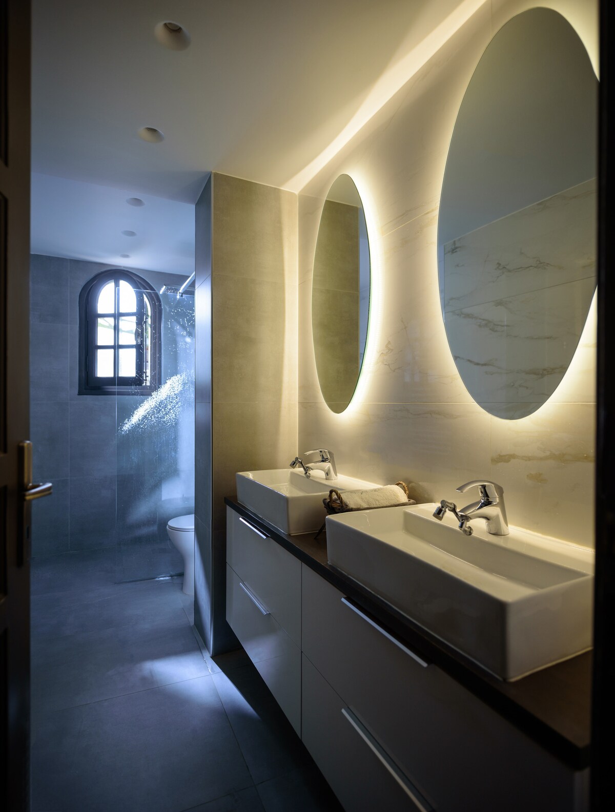 A modern bathroom features two oval mirrors illuminated by soft lighting, enhancing the sleek design. Dual sinks are set on a dark countertop, complemented by cabinetry below. A shower area with glass partition is visible, and natural light filters through an arched window, providing a welcoming atmosphere.