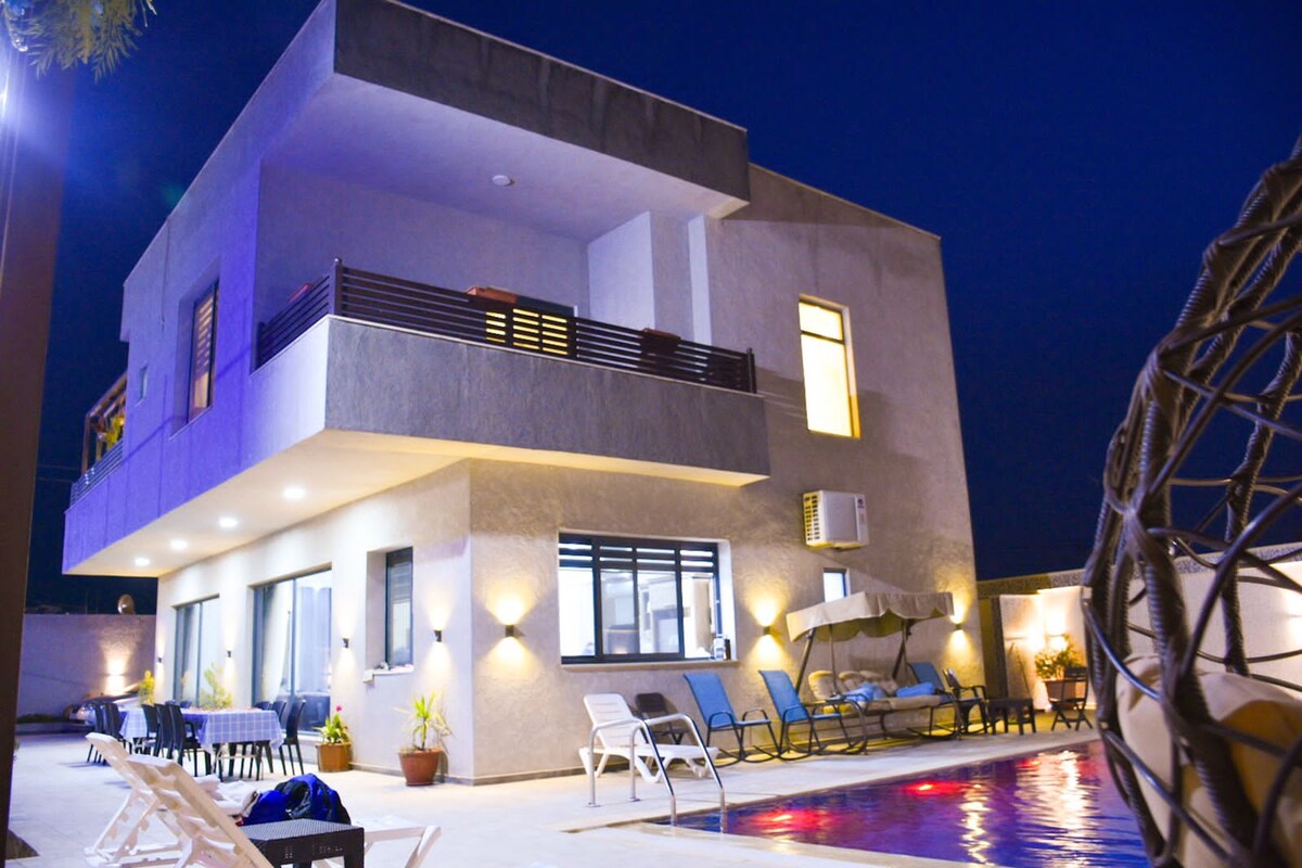 A modern two-story building is illuminated at night, showcasing large windows and a balcony. The outdoor area features a swimming pool surrounded by lounge chairs and a shaded seating area, contributing to a relaxed atmosphere.
