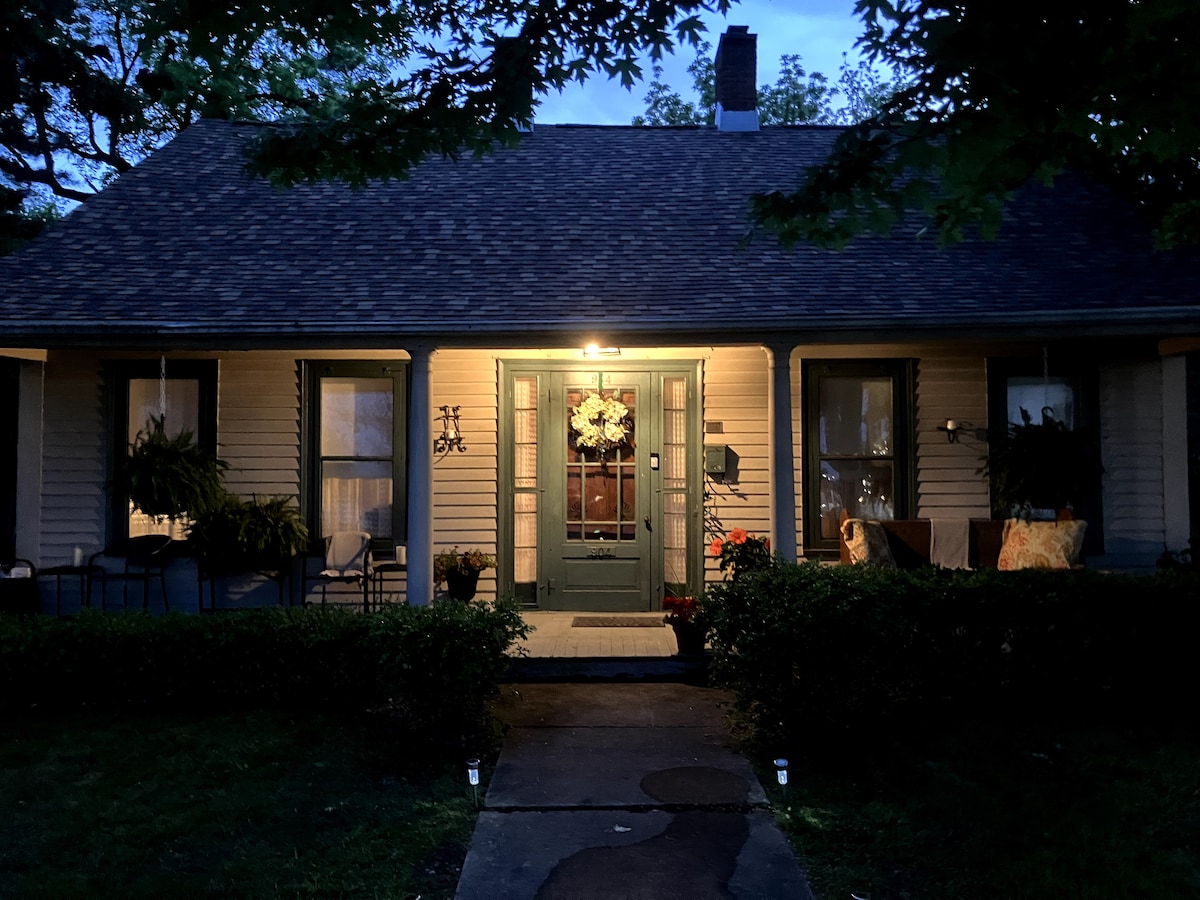 The exterior of a historic home is illuminated by warm outdoor lighting, highlighting a decorative front door with a wreath. Adjacent seating areas are visible on the porch, surrounded by greenery and a neatly trimmed hedge. A stone pathway leads up to the entrance.