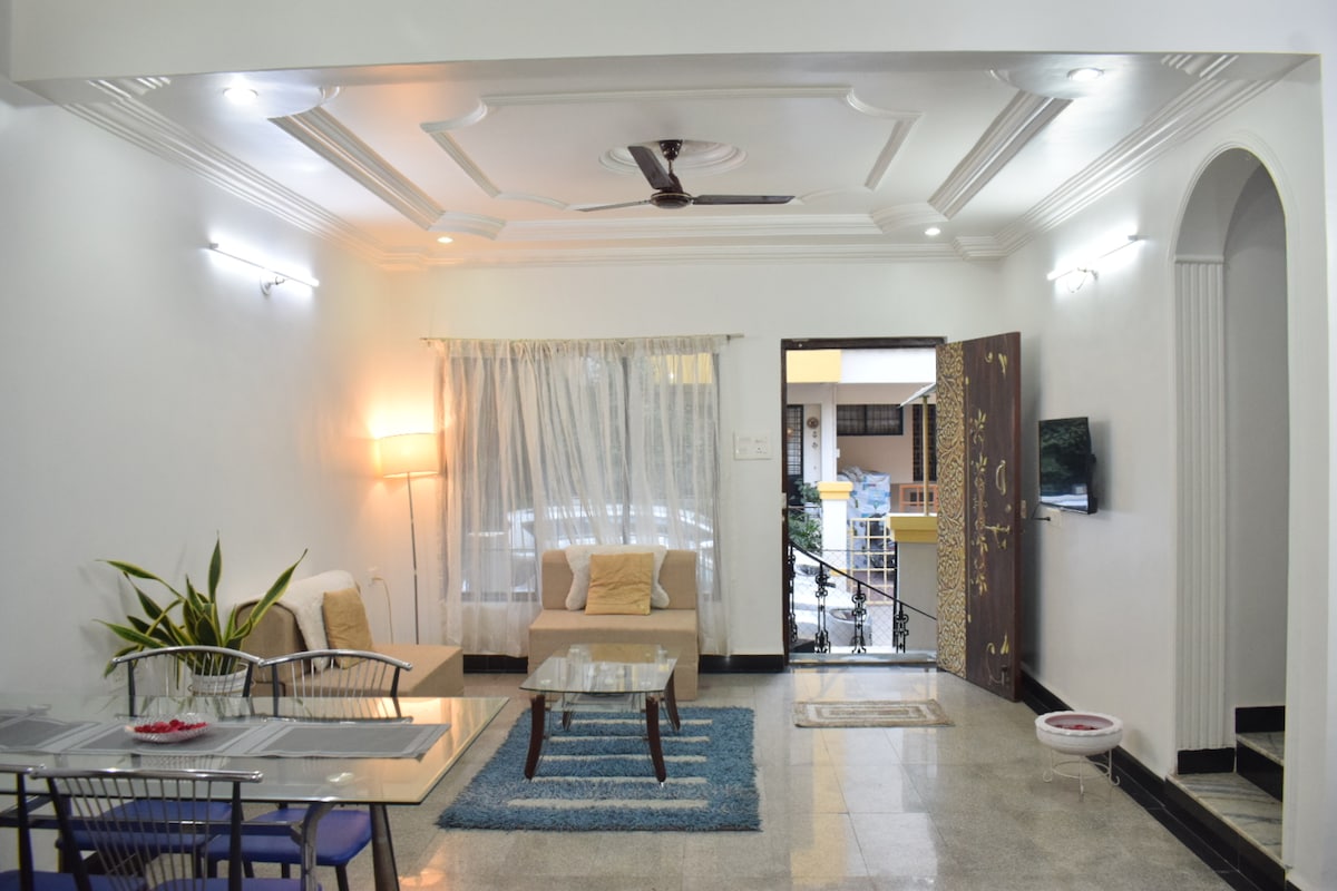A spacious living area is presented, featuring a tasteful arrangement of a comfortable couch and a glass coffee table. Natural light filters through sheer curtains, illuminating the room. An open door leads to an outdoor space, while plants add a touch of greenery to the decor.