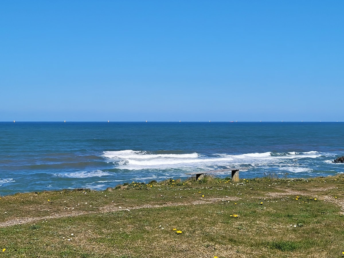 The image captures a clear blue sky above the sea, with gentle waves cresting onto the shoreline. The grassy area in the foreground is dotted with yellow flowers, and distant sailing boats can be seen on the horizon, creating a peaceful coastal scene.