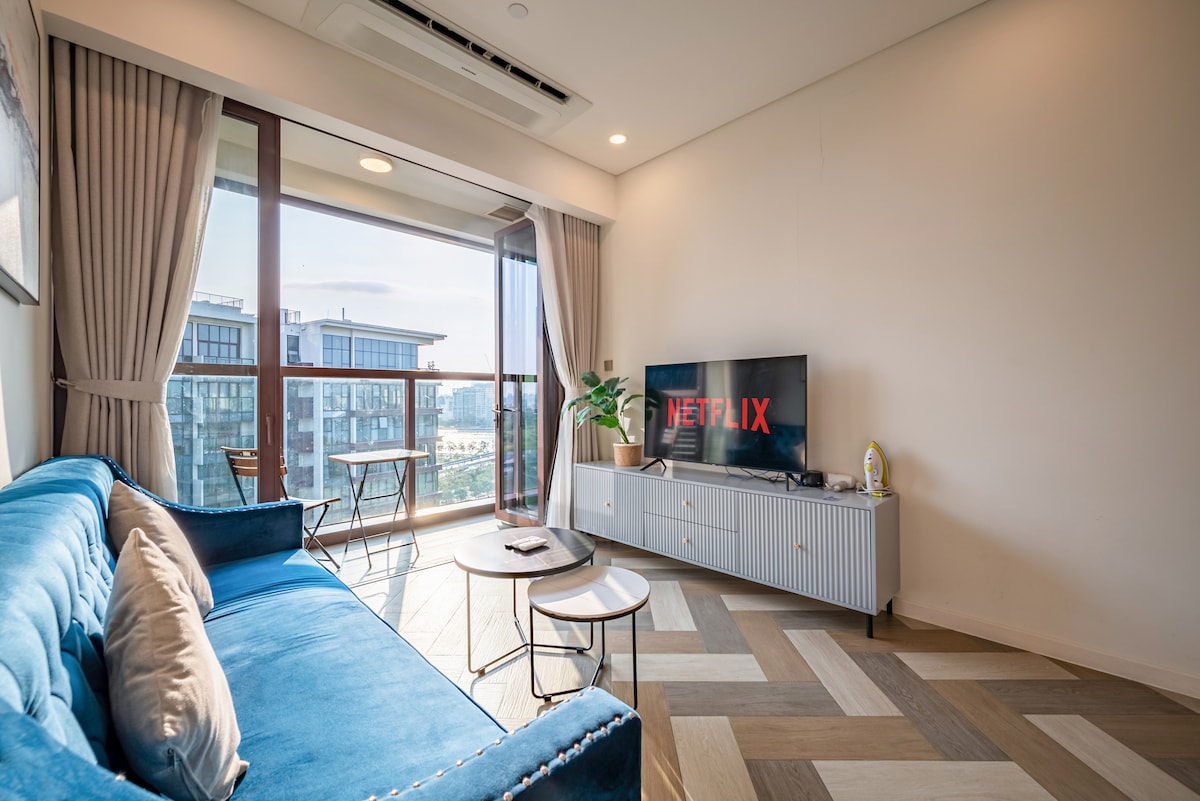 A living room features a comfortable blue sofa paired with a small round table. A large TV displaying Netflix is mounted on a white cabinet. Natural light floods the space through floor-to-ceiling windows, offering views of the outdoor area.