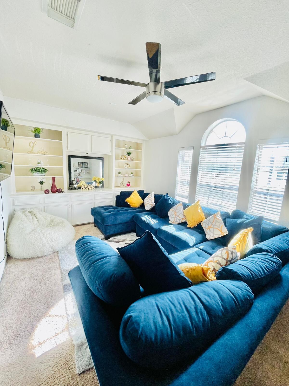 A spacious living room features a large sectional sofa adorned with decorative pillows in a mix of patterns and colors. Natural light filters in through arched windows, illuminating built-in shelving that holds various decorative items. A soft area rug lies beneath a central coffee table.